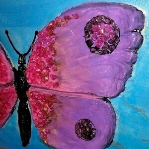 Può includere: Primo piano di una farfalla viola con accenti di glitter rosa sulle ali. La farfalla ha due cerchi neri sulle ali, ognuno riempito di glitter rosa.