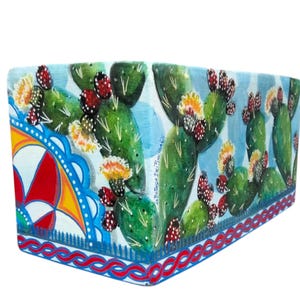 Può includere: Fioriera rettangolare in ceramica con un disegno dipinto a mano raffigurante cactus verdi con fiori rossi e gialli su sfondo azzurro. L'angolo della fioriera presenta un colorato motivo geometrico.