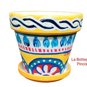 Può includere: Vaso di fiori in ceramica dipinto a mano con sottovaso abbinato. Il vaso presenta una base gialla con motivi decorativi blu, bianchi e rossi. Il bordo ha un design intrecciato bianco e blu. Il testo "La Bottega dei Pincisanti" è visibile.