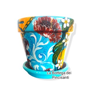 Può includere: Vaso di fiori in ceramica dipinto a mano con sottovaso abbinato. Il vaso presenta un design vibrante con fiori rossi, accenti floreali bianchi e uno sfondo turchese. Il bordo è giallo e arancione. Il testo "La Bottega dei Pincisanti" è visibile.