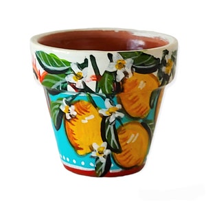 Puede incluir: Una maceta de terracota con un diseño pintado a mano de limones, hojas y flores blancas sobre un fondo turquesa. La maceta tiene un borde blanco y una base roja.