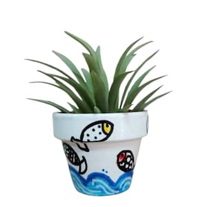 Könnte beinhalten: Kleiner weißer Keramik-Blumentopf mit handgemaltem Ozean-Design. Der Topf zeigt blaue Wellen, schwarz-gelbe Fische und grüne Pflanzenblätter. Der Topf ist etwa 10 cm hoch.