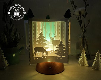 May include: A white papercraft lamp with a winter forest scene. The scene includes two deer, a pine tree, and a snowy background. The lamp is lit and casts a warm glow.