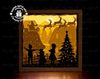 Puede incluir: Una escena navideña recortada en papel con Papá Noel volando en un trineo tirado por renos sobre un pueblo nevado. La escena incluye un muñeco de nieve, dos niños y un árbol de Navidad. La escena está iluminada desde atrás, creando un brillo cálido.