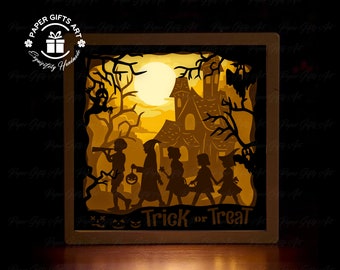 May include: A 3D papercraft light-up Halloween scene with a haunted house, a full moon, and children trick-or-treating. The scene is lit from behind, creating a warm glow. The words "Trick or Treat" are printed at the bottom of the scene.