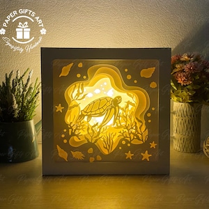 May include: A white wooden box with a 3D paper cut-out design of a sea turtle swimming in the ocean. The box is lit from inside, creating a warm glow. The design features a sea turtle, coral, seaweed, and starfish.