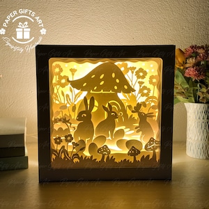 Pode incluir: Uma caixa de madeira branca com um design de papel recortado marrom claro no interior. O design apresenta uma casa de cogumelo com uma pequena porta e janelas, cercada por flores e três coelhos. A caixa é iluminada por dentro, criando um brilho quente.