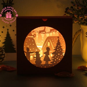 May include: A papercraft light-up Christmas scene with a house, a snowman, a Christmas tree, and two children. The scene is illuminated from behind, creating a warm glow.