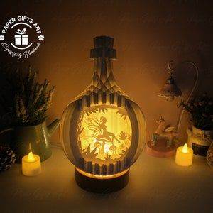 Op de afbeelding: Een witte papieren lamp in de vorm van een fles met een fee en paddenstoelen erin. De lamp is aan en geeft een warm licht. De lamp staat op een tafel met een kleine plant en kaarsen.
