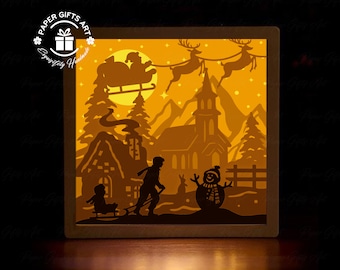 Puede incluir: Una escena navideña iluminada de papel recortado con una iglesia, un muñeco de nieve, una familia y Papá Noel volando en un trineo con renos. La escena está iluminada con luz amarilla cálida.