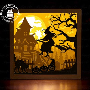 May include: A 3D papercraft light-up box with a Halloween theme. The scene features a witch flying on a broomstick, a haunted house, a black cat, and pumpkins. The box is lit from the inside, creating a warm glow.