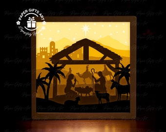 May include: A paper cut-out light-up nativity scene with a yellow and brown color scheme. The scene depicts the birth of Jesus in a stable with Mary, Joseph, and the baby Jesus. There are also palm trees, a donkey, and a sheep in the scene. The scene is illuminated by a warm white light.