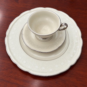 May include: A cream-colored porcelain teacup and saucer set resting on a matching dinner plate. Each piece features a delicate white floral scroll pattern, a smooth center, and a scalloped edge finished with a polished silver metallic trim.