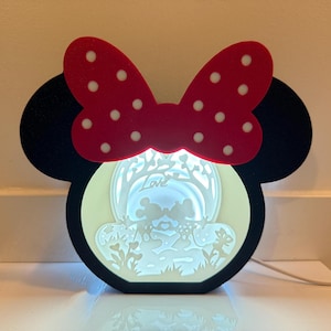 Puede incluir: Una luz nocturna negra y roja con forma de cabeza de Mickey Mouse con un lazo blanco y lunares blancos. La luz presenta un diseño recortado de dos figuras que se toman de la mano con la palabra "Love" encima.