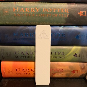 May include: A white bookmark with a dark gray Deathly Hallows symbol on it. The bookmark is placed between a stack of Harry Potter books.