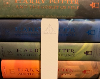 May include: A white bookmark with a dark gray Deathly Hallows symbol on it. The bookmark is placed between a stack of Harry Potter books.