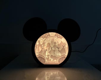 May include: A black Mickey Mouse-shaped lamp with a white silhouette of Disney characters inside. The lamp is lit and the characters are visible through the cutout.