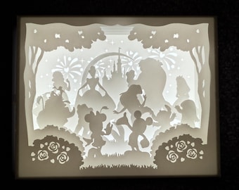 May include: A white papercraft shadow box with a light inside, depicting a scene from a Disney movie. The scene includes a castle, fireworks, trees, and characters like Cinderella, Mickey Mouse, and Minnie Mouse.