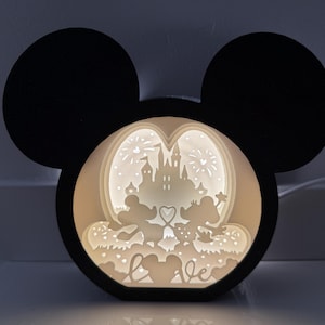 Könnte beinhalten: Eine schwarze Silhouette von Mickeys Kopf mit einer weißen, leuchtenden Szene im Inneren. Die Szene zeigt Mickey und Minnie Mouse vor einem Schloss mit Feuerwerk. Der Text "love" ist am unteren Rand der Szene sichtbar.