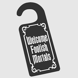 Op de afbeelding: Zwarte deurhanger met een wit versierd frame en de tekst "Welcome Foolish Mortals".