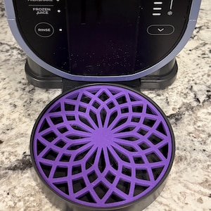 Puede incluir: Una base de licuadora de color morado y negro con un diseño geométrico. La base está diseñada para atrapar las gotas de la licuadora.