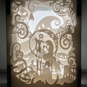 May include: A white papercraft lamp with a silhouette of Jack Skellington and Sally from the Nightmare Before Christmas. The lamp features a moon, pumpkins, and other Halloween-themed imagery.