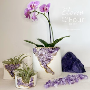 May include: Three white concrete planters with amethyst crystals embedded in the sides. The largest planter holds a pink orchid, while the two smaller planters hold air plants. The planters are on a white surface with loose amethyst crystals scattered around them. The text "Eleven O'Four Decor" and "www.1104decor.com" is visible in the image.