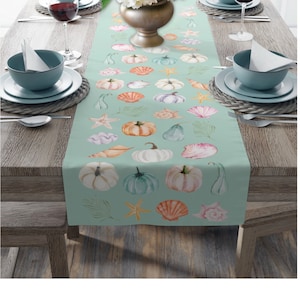 May include: A teal table runner with a pattern of pumpkins, seashells, and starfish. The runner is on a wooden table with two place settings on either side.