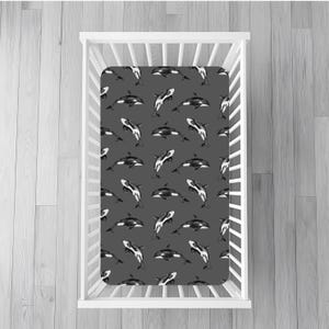 May include: A gray fitted crib sheet with a repeating pattern of black and white orcas. The sheet is inside a white crib, set against a gray wood-paneled background. The orcas are in various positions.