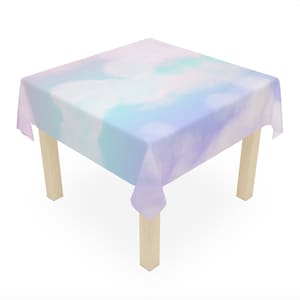 May include: A square tablecloth with a light blue, pink, and purple tie-dye pattern. The tablecloth is draped over a wooden table with four legs.