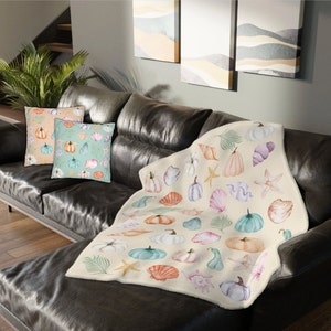Könnte beinhalten: Eine cremefarbene Kuscheldecke mit einem Aquarellmuster aus Kürbissen, Muscheln und Seesternen. Die Decke liegt auf einem braunen Ledersofa mit zwei Kissen, die ein ähnliches Muster aufweisen.