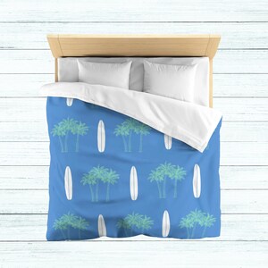 May include: A blue duvet cover with a white surfboard and palm tree pattern. The duvet cover is on a bed with white pillows.