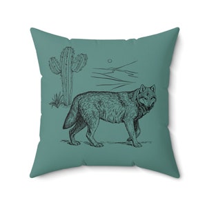May include: A teal blue throw pillow with a black and white illustration of a wolf walking in a desert landscape with a cactus.