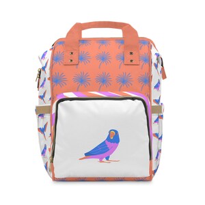 以下が含まれることがあります： ヤシの木の模様と、青、紫、オレンジ色の鳥のイラストが描かれた白いパネルが付いた、コーラル色のバックパック。バッグには上部のハンドル、サイドポケット、ジッパー開閉部が付いています。