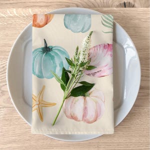 May include: A beige fabric napkin with a watercolour pattern of pumpkins, seashells, and starfish. The pumpkins are in shades of blue, pink, and orange. The seashells are pink and white. The starfish is yellow.