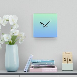 May include: A square clock with a blue and green gradient background. The clock hands are black and the clock is on a white wall. There are books and a wooden calendar block on a shelf below the clock.