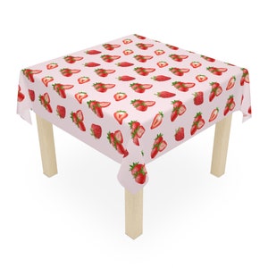 May include: A pink tablecloth with a repeating pattern of red strawberries. The tablecloth is draped over a square wooden table with four legs.