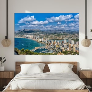 May include: An aerial view of a coastal city with a harbor filled with boats. The city is surrounded by mountains and a blue sky with white clouds.