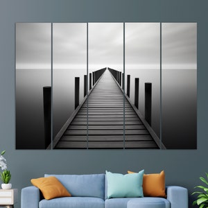 Pode incluir: Uma fotografia a preto e branco de um cais de madeira que se estende para um lago nebuloso. O cais é feito de tábuas de madeira desgastadas e tem postes de madeira de cada lado. A névoa é densa e obscurece o horizonte.