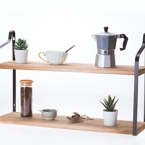 Puede incluir: Estanter&iacute;a de madera de dos niveles con soportes de metal negro. La repisa superior tiene una peque&ntilde;a suculenta en maceta, una taza de t&eacute; blanca y una cafetera plateada. La repisa inferior tiene un frasco de vidrio lleno de granos de caf&eacute;, un peque&ntilde;o taz&oacute;n gris, un peque&ntilde;o ramo de flores secas y una suculenta en maceta.