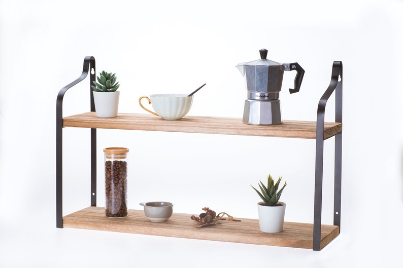 Puede incluir: Estanter&iacute;a de madera de dos niveles con soportes de metal negro. La repisa superior tiene una peque&ntilde;a suculenta en maceta, una taza de t&eacute; blanca y una cafetera plateada. La repisa inferior tiene un frasco de vidrio lleno de granos de caf&eacute;, un peque&ntilde;o taz&oacute;n gris, un peque&ntilde;o ramo de flores secas y una suculenta en maceta.