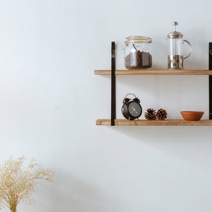 Puede incluir: Una estanter&iacute;a de madera con soportes de metal negro, que contiene un frasco de vidrio de granos de caf&eacute;, una cafetera de prensa francesa plateada, un reloj despertador negro y dos pi&ntilde;as.