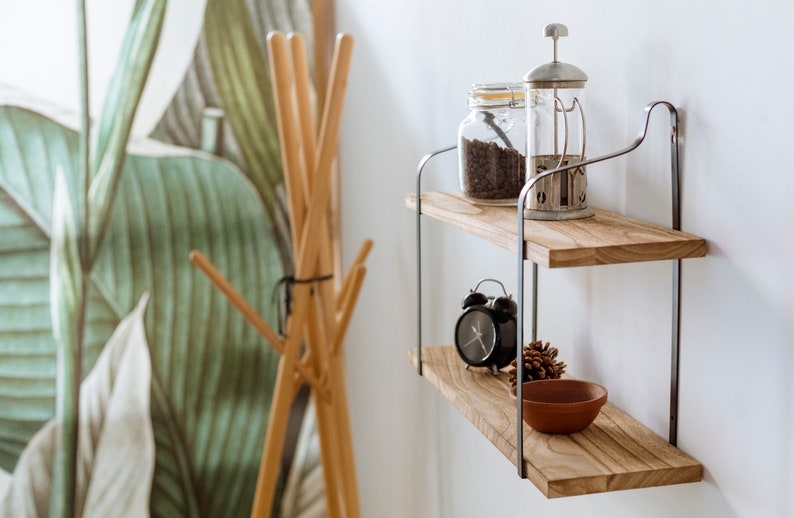 Puede incluir: Estanter&iacute;a de madera de dos niveles con soportes met&aacute;licos, que incluye un tarro de vidrio de granos de caf&eacute;, una prensa francesa, un reloj despertador negro, una pi&ntilde;a y un peque&ntilde;o cuenco de terracota.
