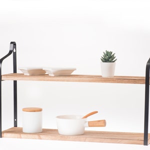 Puede incluir: Una estanter&iacute;a de madera de dos niveles con soportes de metal negro. La estanter&iacute;a est&aacute; decorada con platos blancos, una peque&ntilde;a suculenta en maceta, un tarro de cer&aacute;mica blanca con tapa de madera y una olla de cer&aacute;mica blanca con asa de madera.