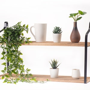 Puede incluir: Una estanter&iacute;a de madera de dos niveles con soportes de metal negro. La repisa superior tiene una taza blanca, una peque&ntilde;a suculenta en maceta y un jarr&oacute;n marr&oacute;n con hojas verdes. La repisa inferior tiene una peque&ntilde;a suculenta en maceta y una jarra blanca.
