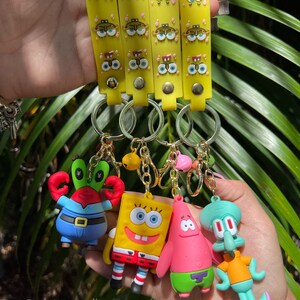 Op de afbeelding: Vier gele sleutelhangers met cartoon gezichten en blauwe ogen. Elke sleutelhanger heeft een ander personage uit de cartoon Spongebob Squarepants: Spongebob, Patrick Star, Squidward Tentacles en Mr. Krabs.