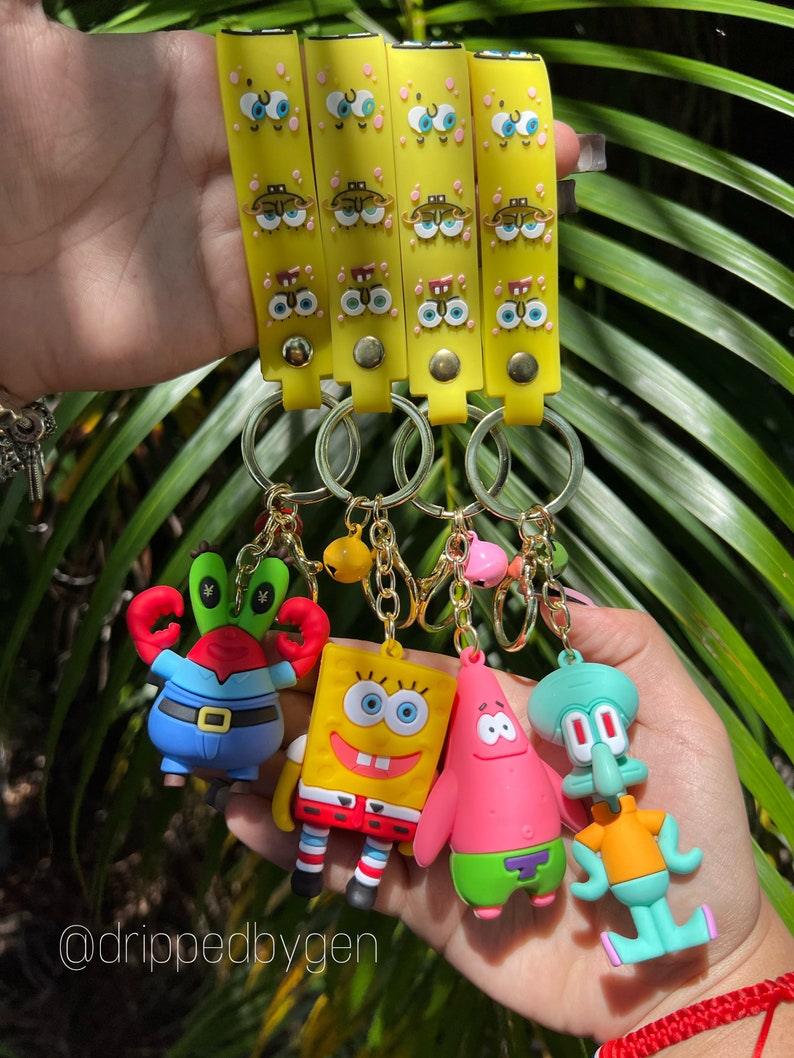 Op de afbeelding: Vier gele sleutelhangers met cartoon gezichten en blauwe ogen. Elke sleutelhanger heeft een ander personage uit de cartoon Spongebob Squarepants: Spongebob, Patrick Star, Squidward Tentacles en Mr. Krabs.