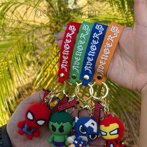 Op de afbeelding: Vier kleurrijke sleutelhangers met het woord "Avengers" erop. Elke sleutelhanger heeft een ander Avenger-personage: Spider-Man, Hulk, Captain America en Iron Man.