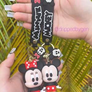 Op de afbeelding: Zwart-witte Mickey Mouse en Minnie Mouse sleutelhanger met rode accenten. De sleutelhanger heeft de woorden "Mickey" en "Minnie" op de bandjes.