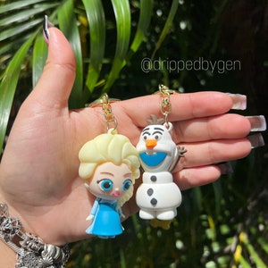Op de afbeelding: Een set van twee Frozen-sleutelhangers met Elsa en Olaf. Elsa draagt een blauwe jurk en heeft blond haar. Olaf is een sneeuwpop met een wortelneus en een blauwe sjaal. De sleutelhangers zijn bevestigd aan gouden kettingen.